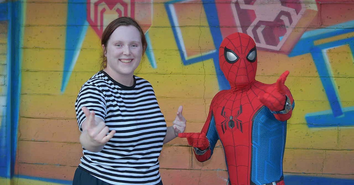 A young, fair-skinned woman posing with Spiderman in front of a brick wall with colourful spider web stencils on it. The young woman is grinning and both are posed with the classic Spiderman web slinging fists.