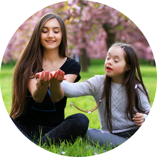 Mum and pre-teen daughter playing together in a park. Both have long dark hair and are smiling at something the mother is holding. The young girl has Down Syndrome. Behind them is a cherry blossom tree full of pink flowers.