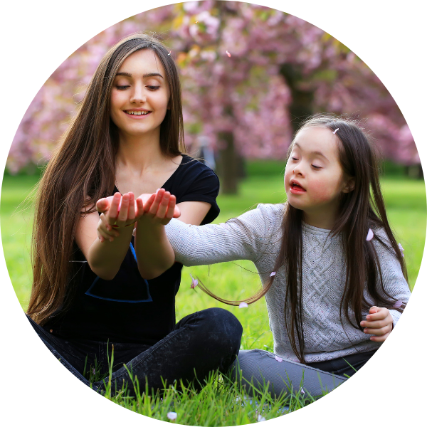 Mum and pre-teen daughter playing together in a park. Both have long dark hair and are smiling at something the mother is holding. The young girl has Down Syndrome. Behind them is a cherry blossom tree full of pink flowers.