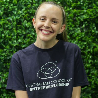 A woman with olive skin and a big smile with her hair tied back in a ponytail, wearing a navy T-Shirt with the logo for the Australian School of Entrepreneurship on it. She is standing in front of a, possibly fake, green hedge.