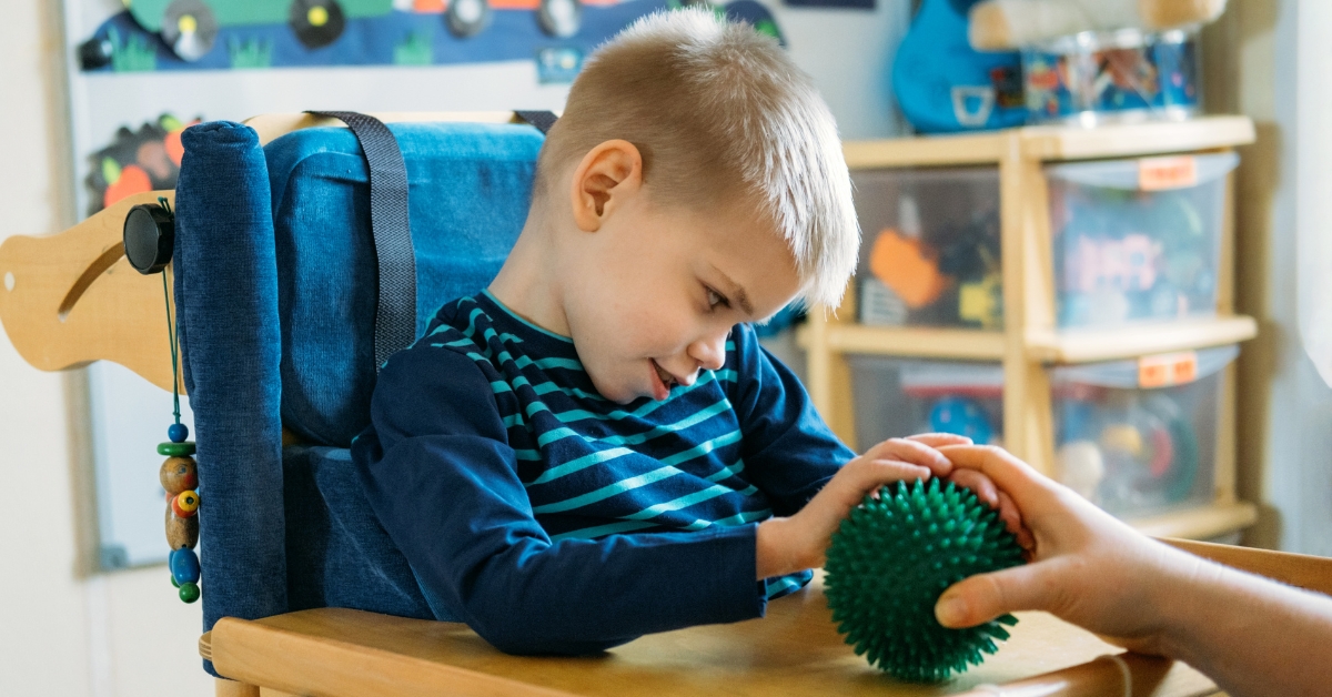A young blond boy in a wooden high chair playing with a spiky green ball sensory toy. Behind him there are boxes of toys and children's artwork depicting cars on a road.