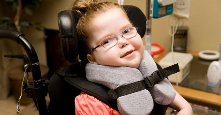 Little girl with glasses wearing a neck brace and using a power wheelchair. Cute as a button and smiling for the camera.