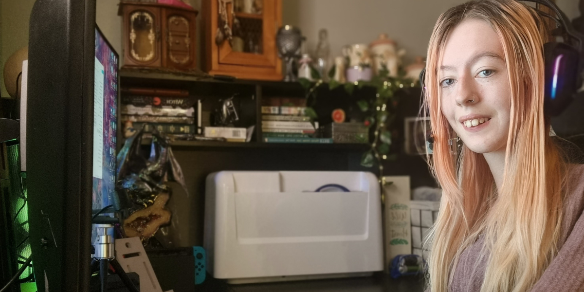 Young person with long fair hair wearing large headphones, sitting in front of a computer screen and smiling at the camera. There is a large printer on the shelf behind her, and the power wheelchair she is using is just visible at the bottom of the frame.