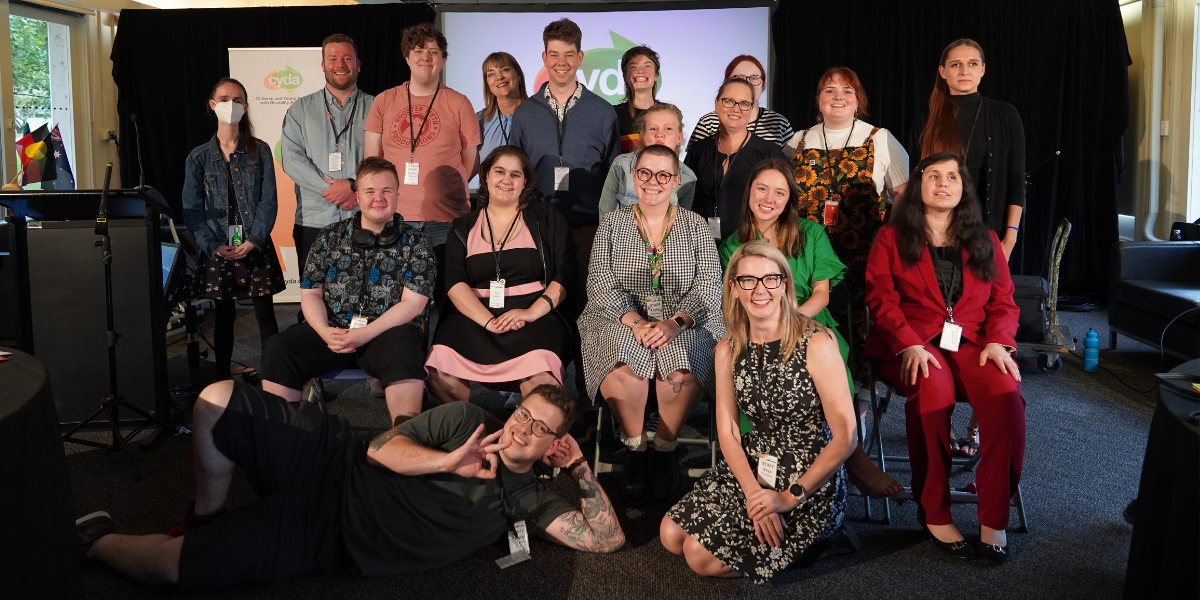 A group of CYDA staff and Youth Council posed together in rows, all smiling for the camera. The man in front is lying on his side making the okay sign. There is signage with CYDA branding behind them.