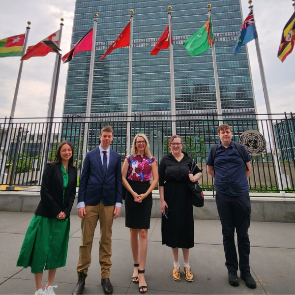 Four young people pictured with CEO of CYDA in front of the UN building in New York. Behind the group are a number of flags flying.
