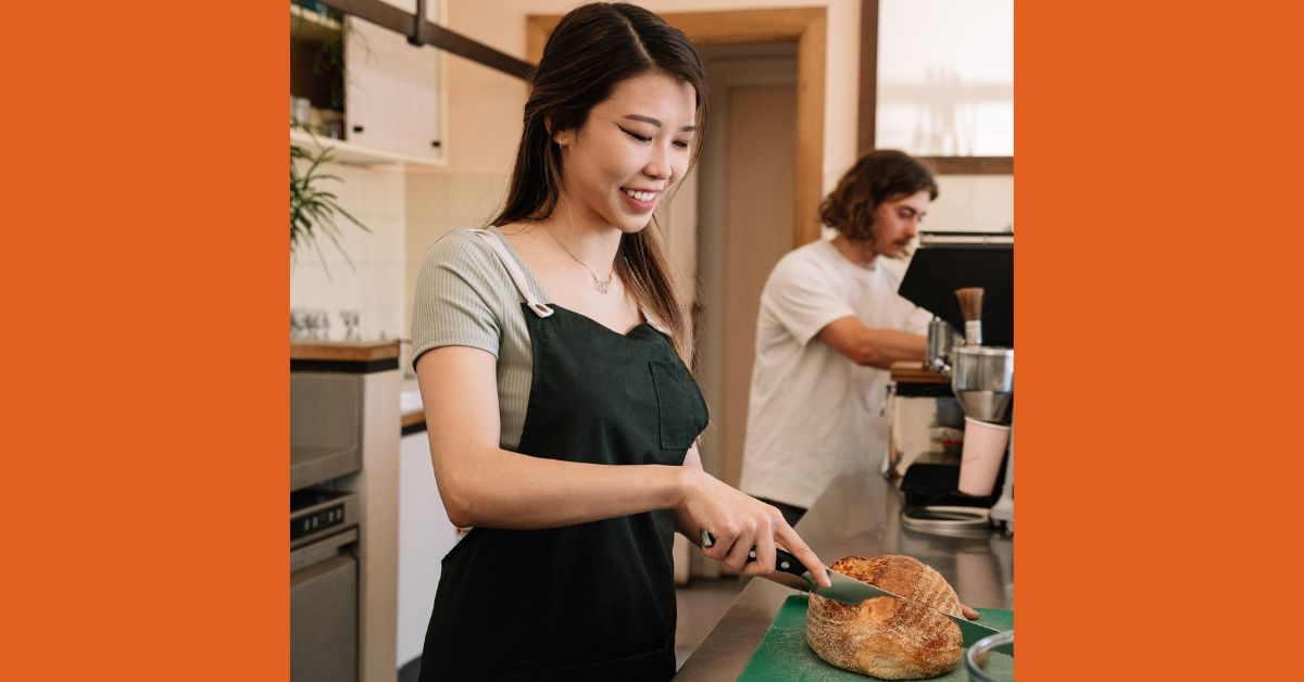 a young person working in a hospitality setting, slicing bread, young person is wearing a black apron and smiling towards the task they are doing.
