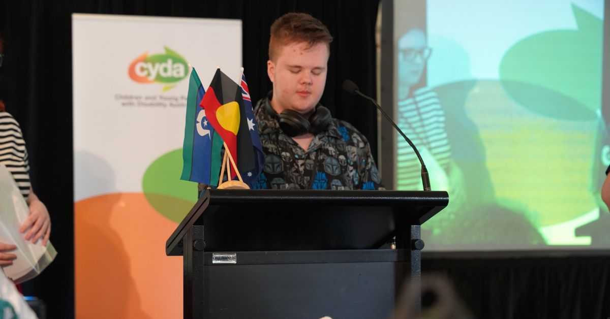 young person presenting and standing behind a lecturn with a microphone one one side and mini flags of first nation, torres strait and australian flags. Young person is wearing black headphones around their neck and a black print shirt. There is a cyda banner in the background.