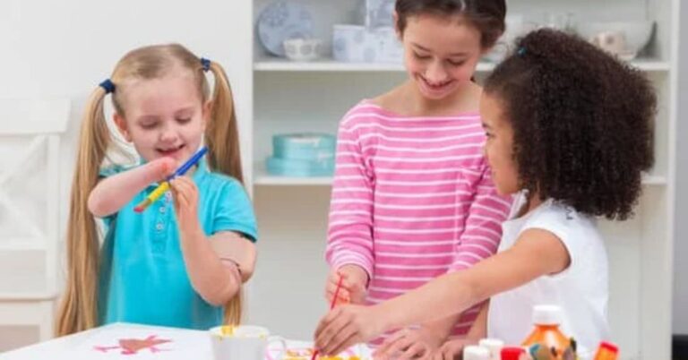 Three school aged children working on art projects. The children are smiling and laughing and dressed in bright coloured clothing. One child has two very long blonde ponytails and is holding a paintbrush with a prosthetic arm.