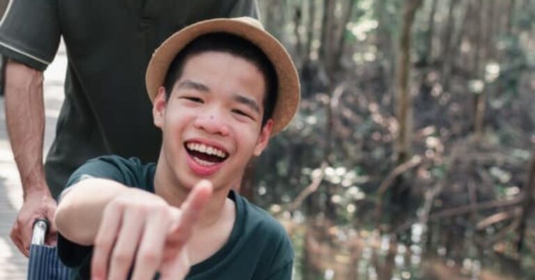 young person smiling and pointing into the camera, wearing a bucket hat with trees in the background.