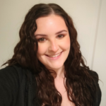 A young, smiling woman with fair skin, big brown eyes and long brown curly hair, wearing a black jacket.