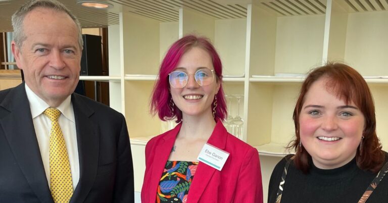 The Honourable Bill Shorten, Minister for the National Disability Insurance Scheme (NDIS), with two young people, Ebe and Kay, standing together, smiling for the camera. Ebe has bright pink hair and wears glasses. Kay has auburn hair. Both are dressed in bright professional attire.