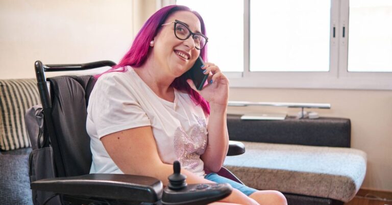 A young woman with long pink hair and black glasses using a motorised wheelchair, chatting on the phone and smiling.