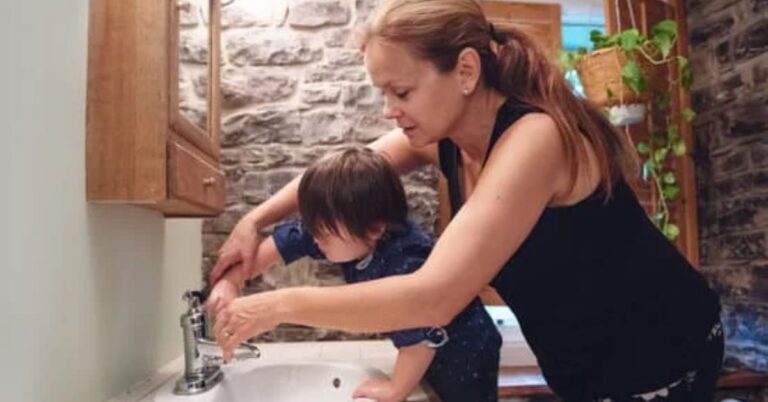 A mum helps a young girl with disability wash her hands in a bathroom sink.