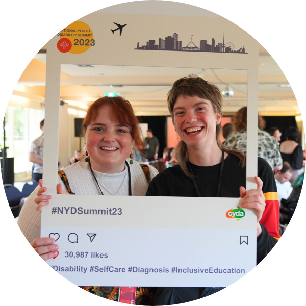 Two young people grinning as they hold up an instagram cutout with with National Youth Disability Summit branding, including a plane and a Canberra cityscape. They are in an event space, and there are people milling about at tables behind them.