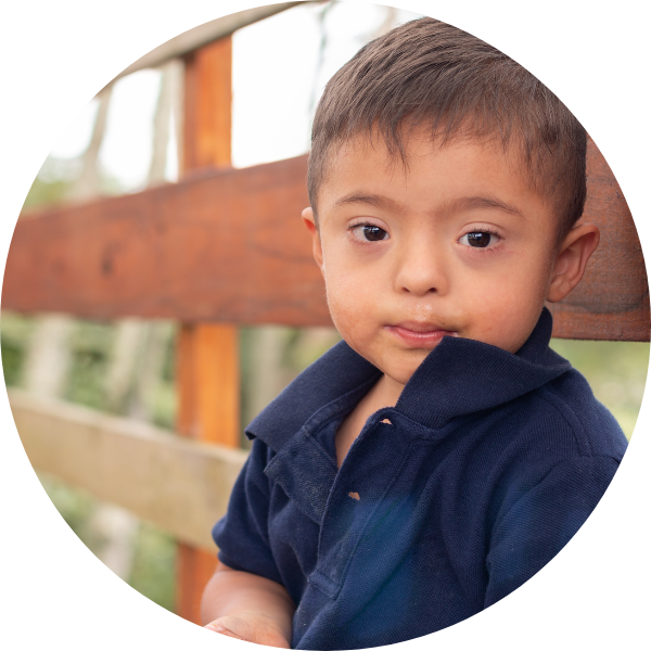 Small boy with Down Syndrome wearing a navy polo shirt and sitting by a wooden fence with greenery behind. He has olive skin, short brown hair and big brown eyes, and is looking into the camera with a pensive expression.
