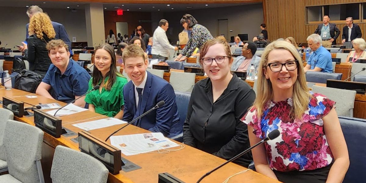 A group of people sitting inside a room at the United Nations in front of desks with microphones, all looking at the camera smiling.