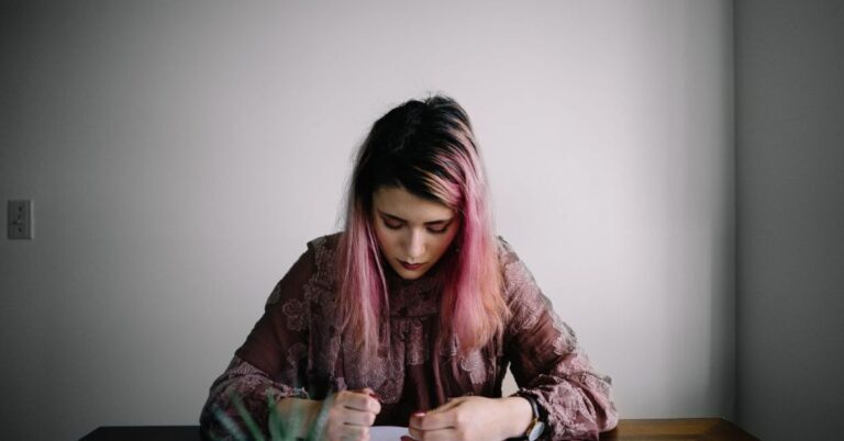 young woman sitting at a desk, looking down towards book, with pink hair.