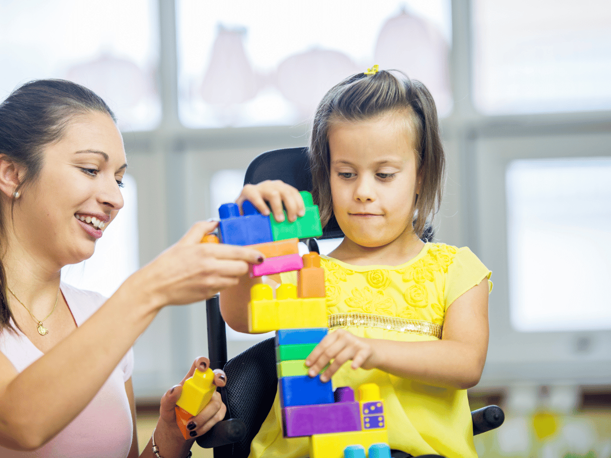 A young person in their wheelchair playing with a stack of colourful blocks. An older person is next to her holding the blocks as well.