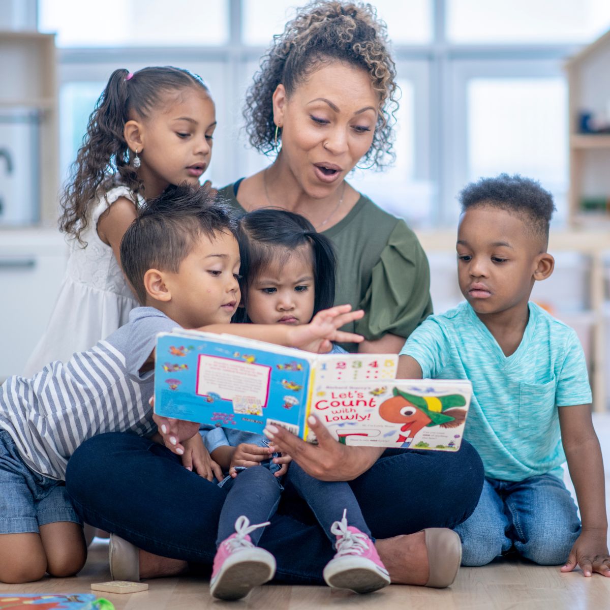 A woman reading a picture book to four small children in a classroom.