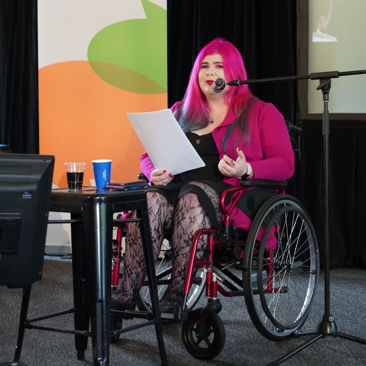 A young woman with bright pink hair, lipstick and jacket. She is on a stage, holding a paper and speaking into a microphone. There is signage behind her with CYDA branding on it. She is using a manual wheelchair.