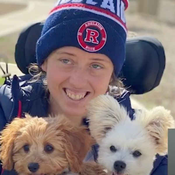 young person smiling and wearing a bright blue and red AFL beanie and holding two small dogs on their lap, sitting in a wheelchair.