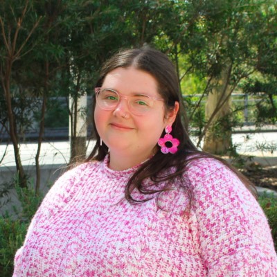 Young person with long dark brown hair, wearing light-framed glasses and bring pink earrings, and smiling into the camera.
