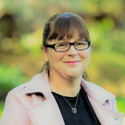 Woman with dark brown fringe and wearing dark-framed glasses. Smiling at the camera with a blurred backrground of greenery.