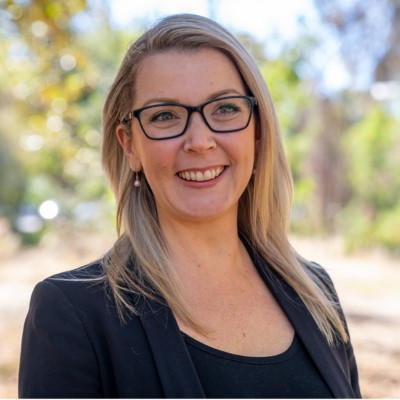 Woman with fair skin and long blonde hair, dark-framed glasses smiling towards the camera with trees blurred in the background.