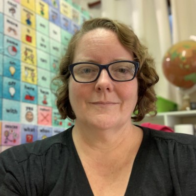 Woman with fair skin and short curly hair, wearing dark-framed glasses and smiling at the camera.