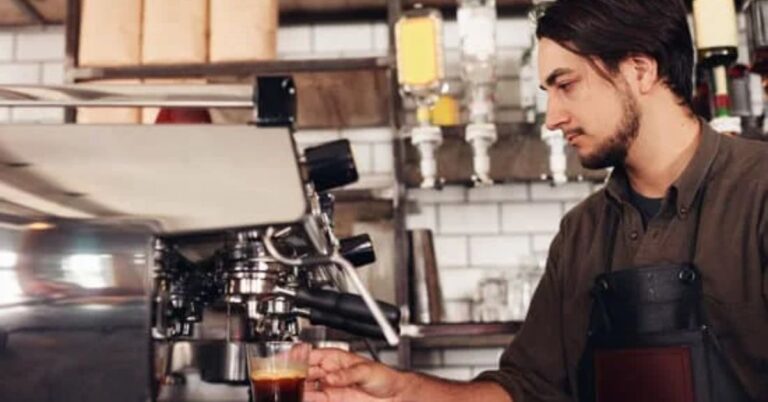 young person with dark hair and a neat, dark beard and moustache, making coffee at a machined.