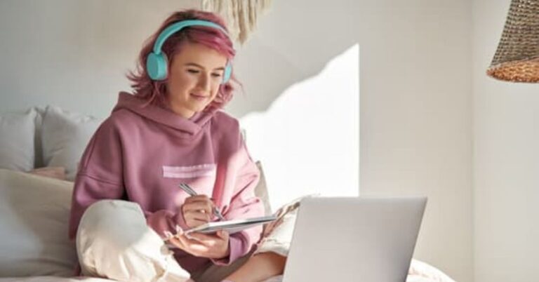 young person with pink shoulder length hair, wearing a pink hoodie, cream jeans and sky blue earphones. Sitting on a bed holding a notebook and looking at a laptop.