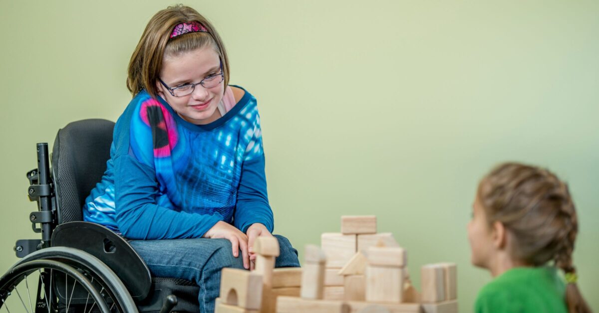 Little girl in a blue top and jeans, wearing a pink headband and using a manual wheelchair. There is a pile wooden building blocks in front of her, and another girl with braided hair and a green t-shirt sitting with her back to the camera. The girls are looking at each other over the blocks and smiling.