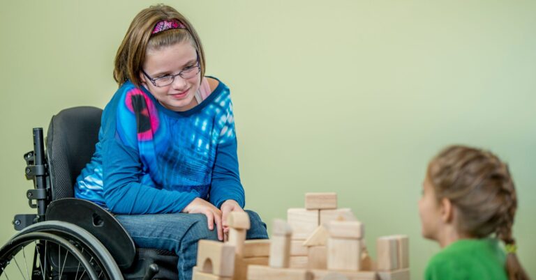 Little girl in a blue top and jeans, wearing a pink headband and using a manual wheelchair. There is a pile wooden building blocks in front of her, and another girl with braided hair and a green t-shirt sitting with her back to the camera. The girls are looking at each other over the blocks and smiling.