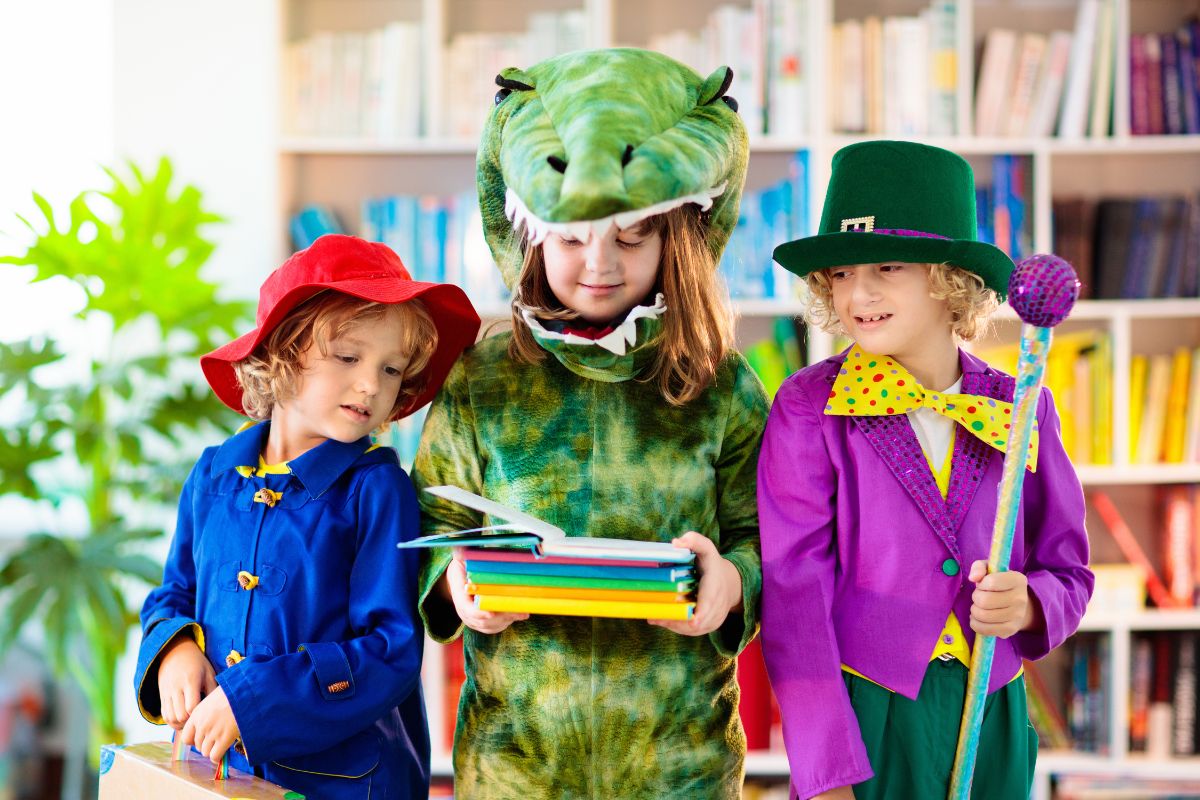 Three little girls in brightly coloured dress up, one in a blue duffel coat and red hat, one as a dinosaur, one in a green and purple top hat and coat with a yellow bowtie. Dinosaur girl is in the middle holding a stack or colourful books which they are all looking at eagerly.