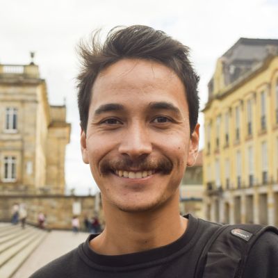 Man smiling into the camera, with short dark hair and a moustache. In the background there are old buildings.