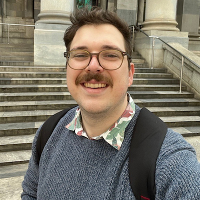 A young, fair-skinned man with dark hair, glasses and a moustache, wearing a grey knit jumper, collared shirt and a backpack. He's taking a smiling selfie in front of an old building with many concrete steps.