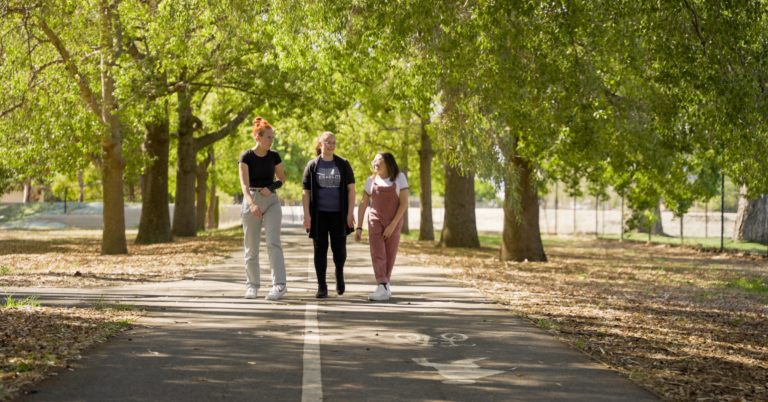 A photograph shows three young people walking through a sunny park under green foliage.