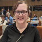 A young smiling woman with fair skin, glasses and hair back from her face, wearing a black shirt. She is in a large conference space with rows of people at desks behind her and a microphone on the desk before her.