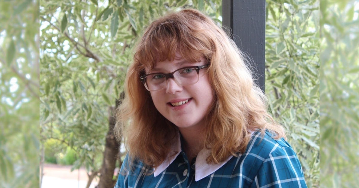 A smiling girl with glasses, fair skin, thick blond hair and a fringe. She is standing on a porch, wearing a school uniform. There are gum trees in the background.