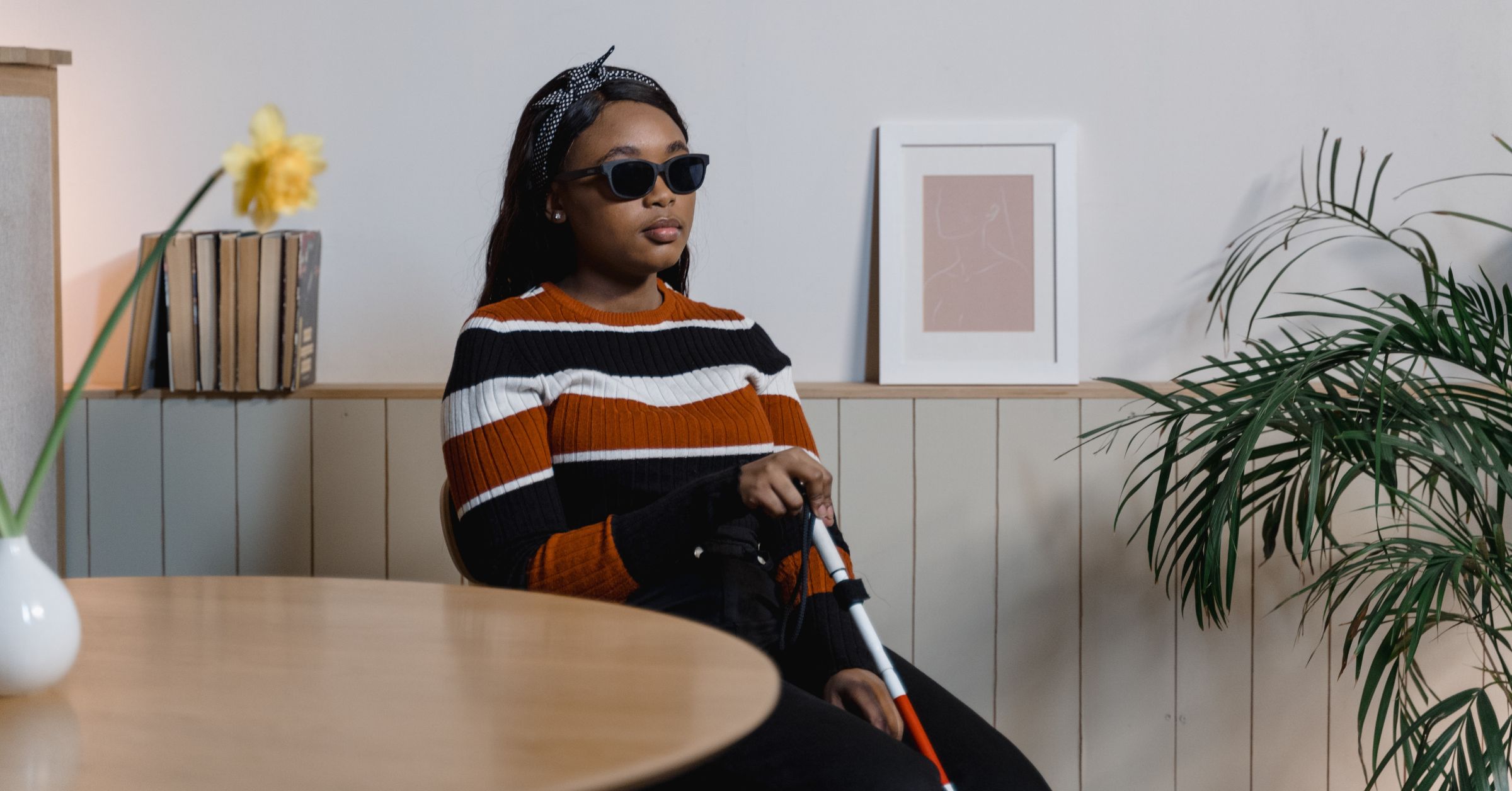 A young black woman wearing sunglasses and a black, brown and white striped wool jumper, sitting by a table holding a white cane. There are pot plants dotted around and an indistinct, framed pink picture on the wall.