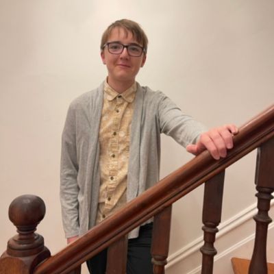 A young man standing on a set of stairs holding the handrail with one hand. He has brown hair with pale skin and dark rimmed glasses. He is wearing dark trousers, yellow patterned button up shirt and a grey cardigan. He has a closed mouth smile.