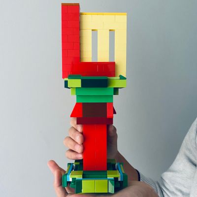 A hand holding trophy represented the Lego Master symbol with Letter L and M design. The trophy has a green brick base with a red brick post. L and M letters made by red and yellow colour bricks are situated on the top of the post.