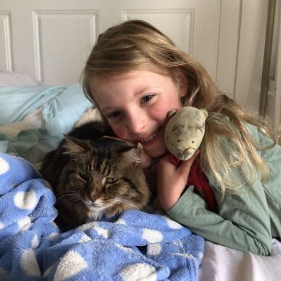 A photo of a young pale faced girl with long blond, brown hair hugging a well-loved teddy and a brown cat. The cat looks serious, lying on a blue blanket with white spots.