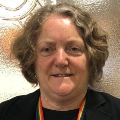 A smiling, fair skinned, older woman with short wavy grey hair wearing a rainbow lanyard.