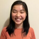 Young femme presenting Asian person, smiling brightly into the camera, with shoulder length straight black hair, wearing an orange jumper.