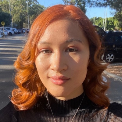 A young femme presenting person with curled orange/red hair smiling slightly. Behind them is a tree-lined suburban street.