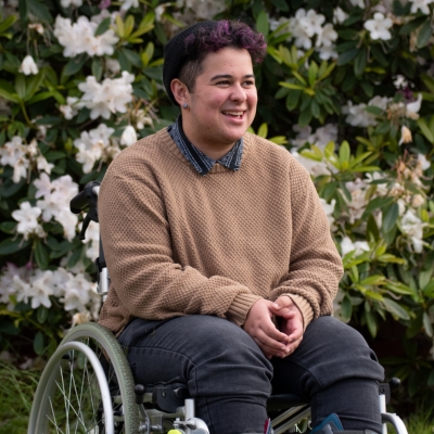 A young man using a manual wheelchair sitting outside with flowers behind him. He is wearing a beige jumper with a collared shirt and has his hands clasped in front of him.