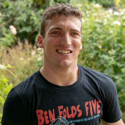A smiling man with fair skin, short, curly, brown hair, and a square jaw standing outside in a garden, wearing a Ben Folds Five band t-shirt.