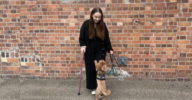 A young woman with fair skin and long, dark hair, wearing all black and holding a red walking stick, standing in front of a red brick wall. She is looking down at a small dog whose lead she is holding. The dog is jumping at her leg for attention.