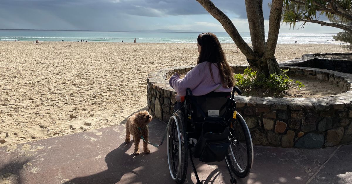 A young woman with long dark hair sitting in a manual wheelchair. She is wearing a purple jumper and has a small assistance dog beside her. She is looking out at a sandy beach with a blue sky.